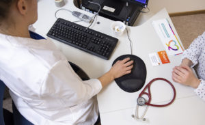 Kuvassa lääkäri käyttää tietokonetta ja potilas odottaa vieressä. Pöydällä on stetoskooppi.