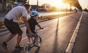 Kuvassa vanhempi henkilö auttaa pientä lasta ajamaan polkupyörällä. Molemmat ovat selin ja suuntaavaat ilta-aurinkoon päin.