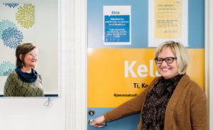 Sari Kenttä avaamassa Kelan toimipisteen ovea Haukiputaan hyvinvointikeskuksessa. Viereisessä peilissä Sari Hedemäen kasvot, molemmat hymyilevät toisilleen yhteisen peilin kautta.