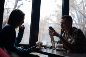Kaksi ihmistä kahvilassa, toisella puhelin kädessä, ikkunasta näkyy syksyinen maisema.